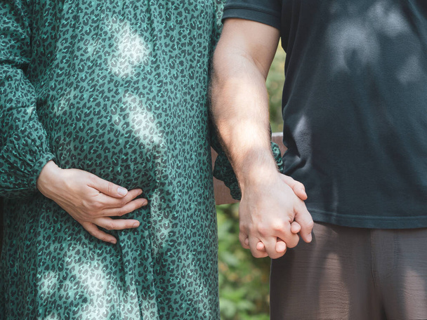 Man and woman hold hands in nature on a sunny day. The woman is pregnant. A pregnant woman is dressed in a green dress, supporting her belly with one hand. Pregnancy and motherhood concept. Close-up. - Photo, Image
