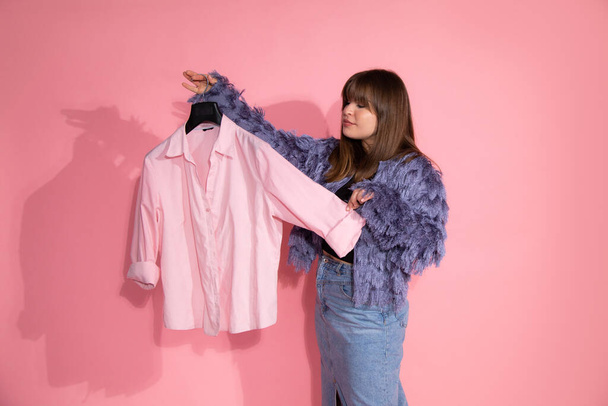 молодая женщина стилист держит вешалку с рубашкой на розовом фоне в студии. Основная концепция гардероба. Блогер преподает классы по созданию стильного образа. Концепция фото - Фото, изображение