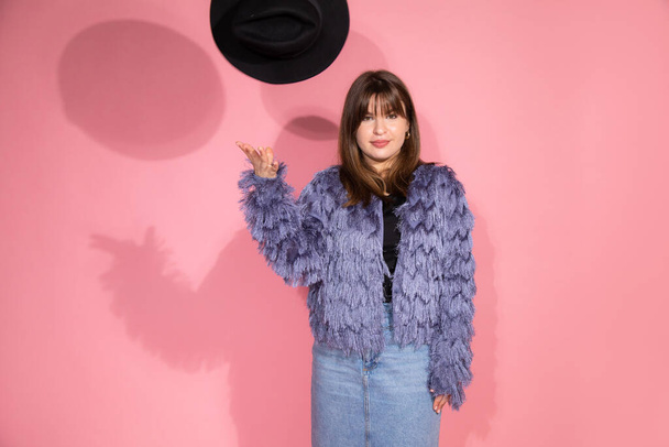 Photo of young, beautiful dark-haired woman in fluffy same hat made of faux fur. With black hat looks into frame, play with shadows and light. Pink background. studio shot. - Fotó, kép