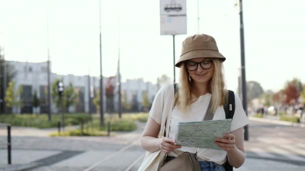 Молодая туристическая женщина с рюкзаком ищет правильное направление на карте ждет общественного транспорта на трамвайной станции на открытом воздухе. Путешествие в Европу - Кадры, видео