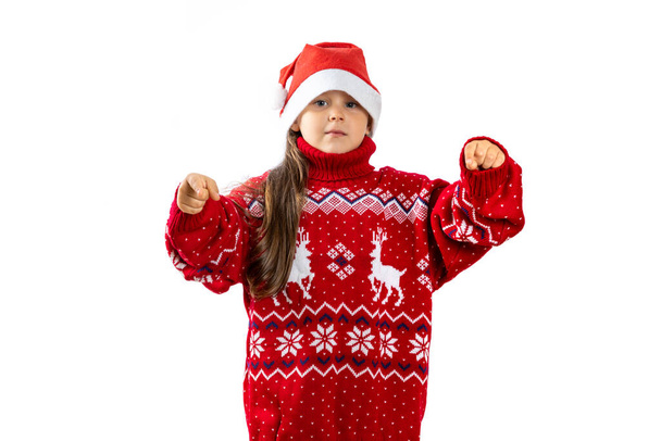 portrait of six-year-old girl in red Santa Claus hat in red knitted Christmas sweater with reindeer pointing fingers at camera, isolated on white background - Фото, изображение