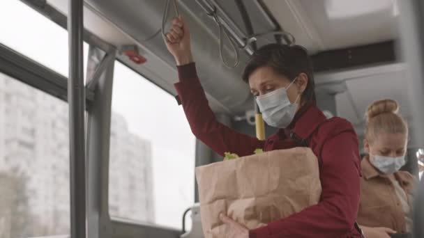 Bajo ángulo de mujer caucásica de pelo corto con máscara y gabardina roja, bolsa de papel con comestibles, autobús - Imágenes, Vídeo
