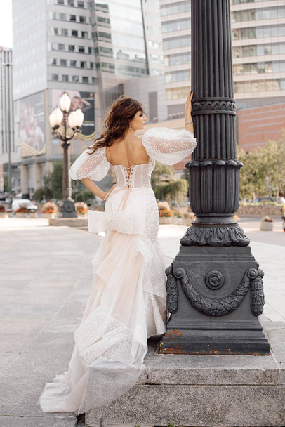 Фото нареченої з темним волоссям у білій весільній сукні на лампочці на тлі міських будівель, виглядає щасливо. Щасливі миті. Вид спереду. Нерозпізнана людина
 - Фото, зображення