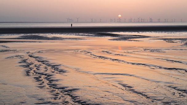 One of the one hundred Iron Men statues (which make up Antony Gormley's Another Place art installation) seen at the end of muddy channels on Crosby Beach near Liverpool at sunset in April 2021. - Photo, Image