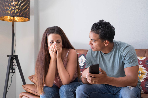 Freund bittet um Erklärung über Handy-Nachrichten seiner Freundin, die auf einer Couch im Wohnzimmer in einem Haus mit dunklem Hintergrund sitzt - Foto, Bild