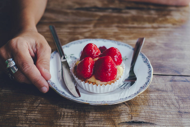 Κοντινό πλάνο του χεριού φράουλα έκανε νόστιμο μικρό κέικ και το χέρι κρατώντας το πιάτο σε ένα ξύλινο τραπέζι. Έννοια των ανθρώπων και της ζάχαρης φυσικά φρούτα και τρόφιμα - Φωτογραφία, εικόνα