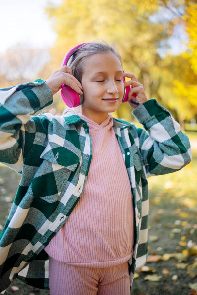 Adorable teenage girl listening to music in big pink headphones in autumn park. - Photo, Image
