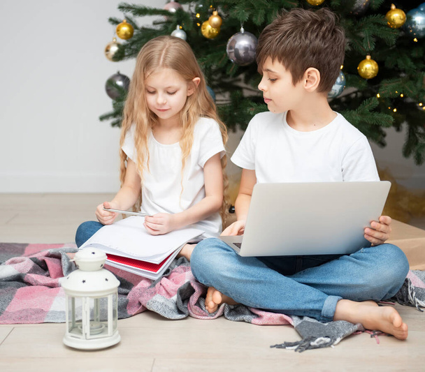 Arbre de Noël dans un grand salon. Les enfants jouent près du sapin de Noël. Un garçon avec un ordinateur portable communique avec des amis. - Photo, image