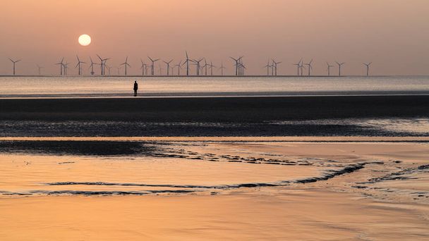 Цветной солнечный свет отражается от ила на пляже Кросби на закате в апреле 2021 года. Он является одной из ста статуй Железных людей, которые составляют художественную инсталляцию Антония Гормли "Другое место", захваченную недалеко от Ливерпуля.. - Фото, изображение