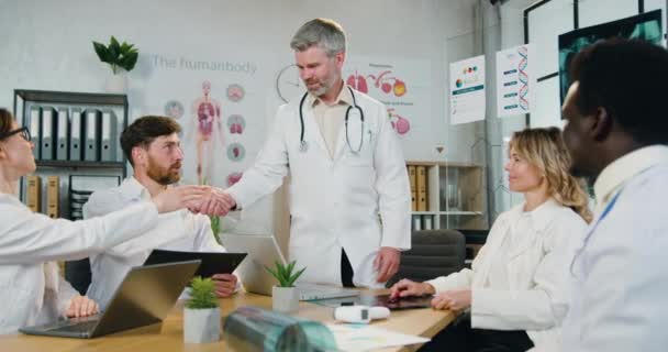 Workflow in medical office where handsome confident qualified bearded doctor greeting every member of his multiethnic team with handshakes before the start of joint conference - Materiał filmowy, wideo