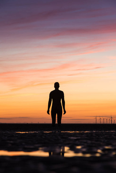 An Iron Man watches the sun setting behind the spinning windmills of Burbobank Windfarm off the Sefton Coast in Liverpool.  He is one of the 100 Iron Men statues created by Antony Gormley which occupy this stretch of Crosby beach near Liverpool. - Foto, Imagem