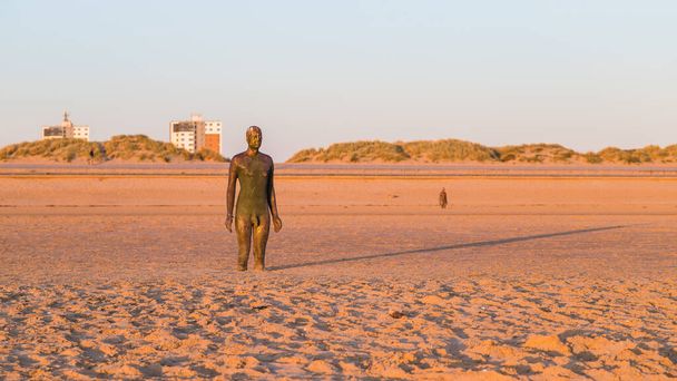 Длинная тень позади Железного человека на закате на пляже Кросби в апреле 2021 года. Это, и то, что на заднем плане часть другого места, художественная инсталляция возле Ливерпуля, состоящая из 100 статуй Железных Людей, созданных Антонием Гормли. - Фото, изображение