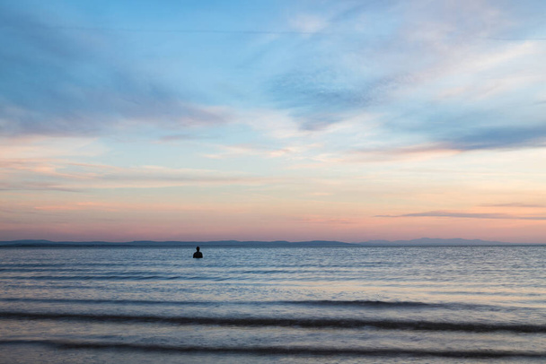 Samotny Iron Man widziany na plaży w Crosby podczas przypływu o zmierzchu w kwietniu 2021. Jest częścią Innego Miejsca, instalacji artystycznej na plaży Crosby koło Liverpoolu złożonej ze 100 posągów Iron Mana stworzonych przez Antoniego Gormleya. - Zdjęcie, obraz