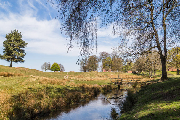 Eine hölzerne Fußgängerbrücke überspannt einen kleinen Bach in der Landschaft von Cheshire im April 2021. - Foto, Bild