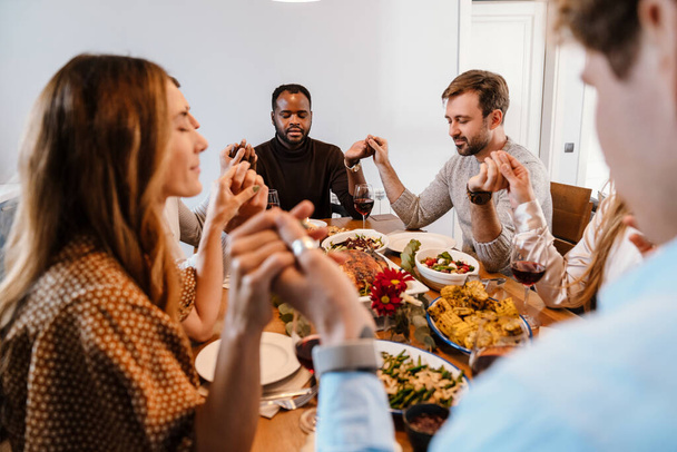Multiracial friends holding hands and praying during thanksgiving dinner at home - Photo, Image
