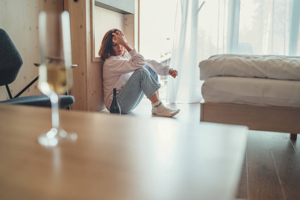 Sad woman sitting on the floor next to window in the bedroom with an opened bottle of alcohol. Unfocused white wine glass on the foreground table. Mental health and alcoholism problems concept image. - Fotó, kép