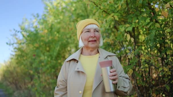 Bela loira de cabelos idosos em roupas elegantes bebe uma bebida quente de café, desfrutando da natureza e caminhando no parque de outono - Filmagem, Vídeo