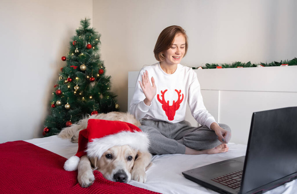 Imádnivaló lány laptoppal és kutyával karácsonyi hangulatban. Szabadúszó otthonról dolgozik az új évben. Egy lány egy golden retriever egy Mikulás kalap kommunikál videó linken keresztül egy laptop. - Fotó, kép
