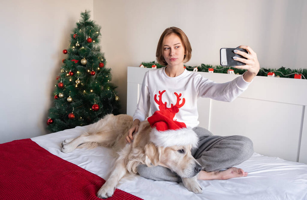 Portre of a young woman with a dog in a Christmas atmosphere. The girl looks at the phone in the hat of Santa Claus with a golden retriever on the bed near the Christmas tree. - Valokuva, kuva