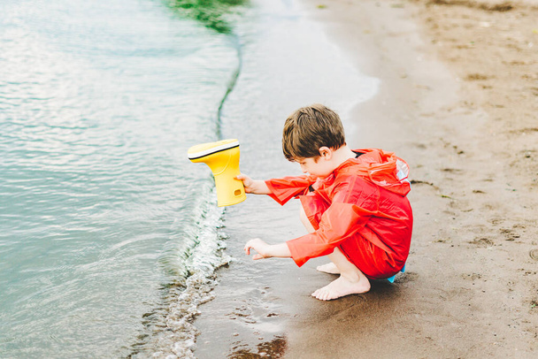 Jongen in een rode regenjas giet water uit gele rubberen laars in het meer. Kind speelt met water in de vijver. Kind heeft plezier buiten in de buurt van Riverside - Foto, afbeelding