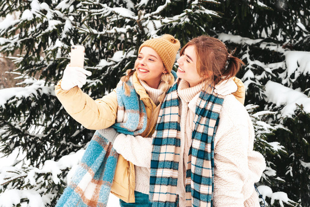 Two young beautiful smiling hipster female in trendy warm clothes and scarfs.Carefree women posing in the street in park. Positive pure models having fun in snow. Enjoying winter moments.Taking selfie - Φωτογραφία, εικόνα