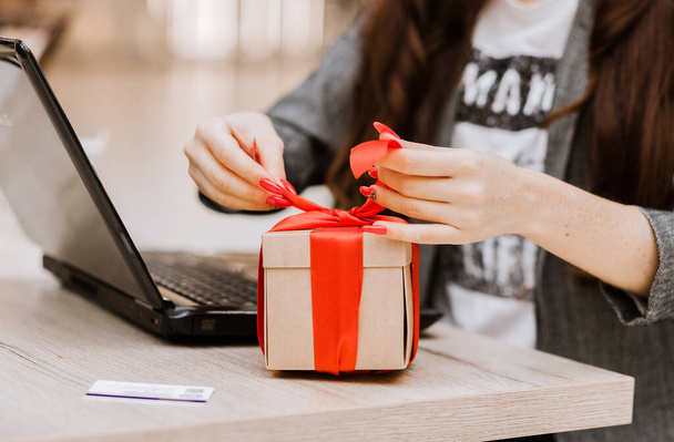 The woman opens the gift. There is a laptop and a credit card on the table. Online shopping and gift concept. Horizontal photo - Foto, Imagem