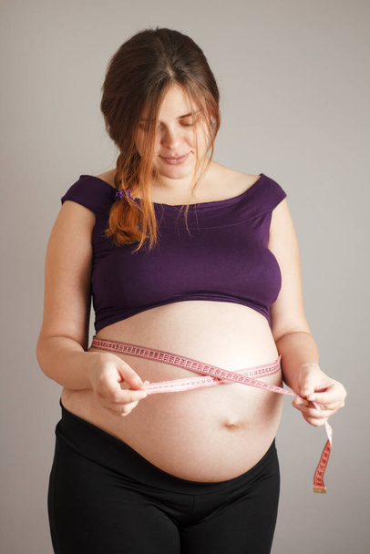 mooie roodharige zwangere vrouw gebruik tape meten op grote buik op grijze studio achtergrond, vrouwelijke lichaam figuur controle tijdens de zwangerschap - Foto, afbeelding