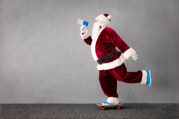 Good news! Santa Claus riding skateboard. Portrait of senior man wearing Christmas costume. Santa Claus shouting by loudspeaker. Xmas holiday concept - Photo, Image