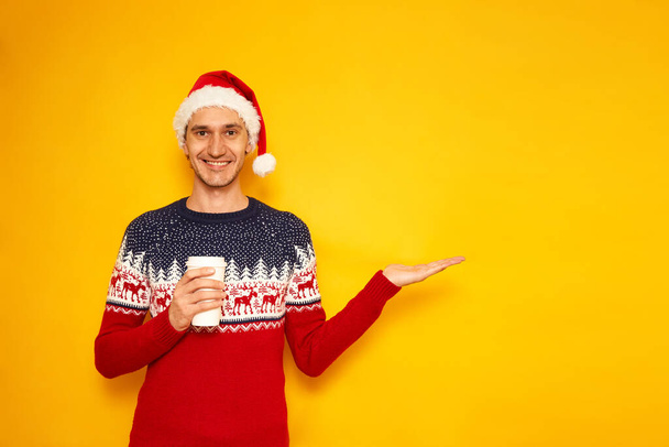 Sıcak içecekler için kağıt bardaklı neşeli adam Noel Baba 'nın süveteri Noel Baba şapkası boş bir reklam alanına işaret ediyor sarı arka planda mesaj konsepti için yer var - günaydın - Fotoğraf, Görsel