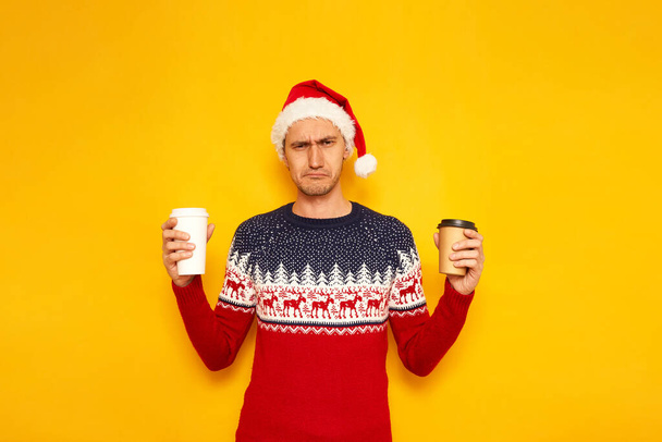 Сумний, незадоволений, розчарований чоловік у різдвяному светрі з північнооленячим червоним капелюхом Санта Клауса тримає паперові чашки з гарячим чаєм, кавою. Жовтий фон з простором для текстової концепції нового року - Фото, зображення