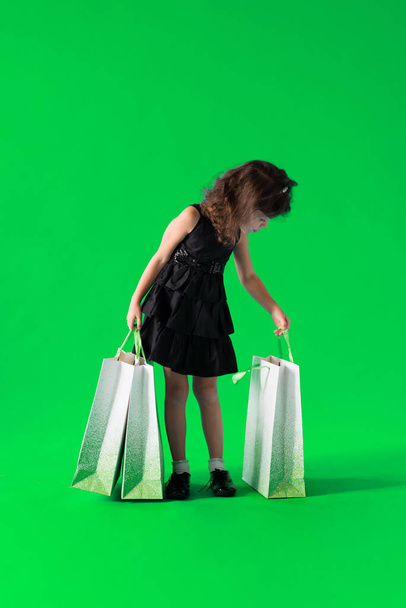 Girl in a black dress with paper bags goes shopping for discounts on a green background - Φωτογραφία, εικόνα