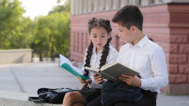 Kinder zwei Schulkinder hispanische kleine Schülerin und Junge Schülerin mit Buch sitzt draußen Kind öffnet Tagebuch liest Bemerkung für schlechtes Benehmen wirft Notizbuch beleidigt lenkt Kreuze Arme - Filmmaterial, Video