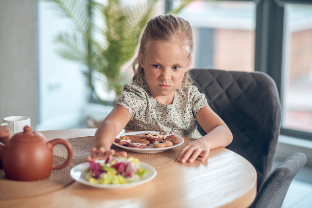 A small girl looking unpleased while looking at the food on the table - Zdjęcie, obraz
