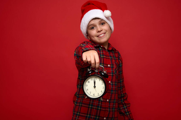 Lächelnder süßer Junge mit Weihnachtsmütze und rot kariertem Hemd hält in ausgestreckten Händen einen schwarzen Wecker mit Mitternacht auf dem Zifferblatt und zeigt ihn in die Kamera. Weihnachts- und Neujahrskonzept für Werbung - Foto, Bild