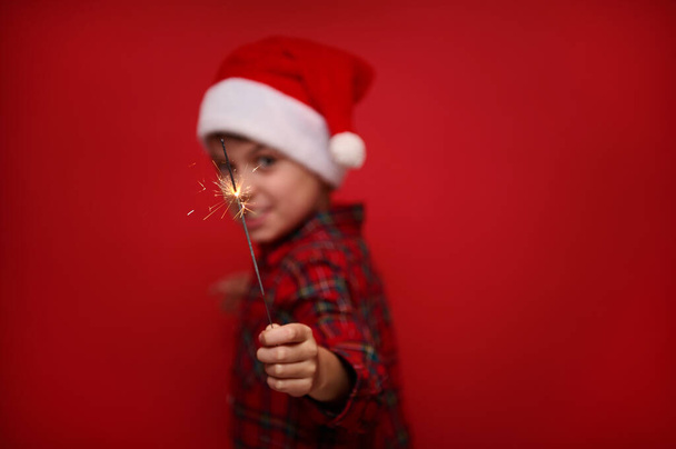Concentre-se nos brilhos de fogo nas luzes de bengala sparklers na mão de menino bonito borrado em chapéu de Papai Noel e camisa quadriculada, isolado sobre fundo de cor vermelha com espaço de cópia para anúncio de Natal - Foto, Imagem