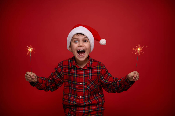 Conceito de celebração de Natal e Ano Novo para propaganda. Engraçado incrível menino bonito em chapéu de Santa com sparklers contra fundo de cor vermelha com espaço de cópia - Foto, Imagem