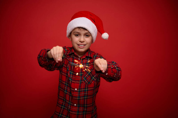 Handsome adorable preteen boy, beautiful child in Santa Claus hat and checkered shirt enjoys the Christmas party, plays with Bengal lights, sparklers, isolated o red background with copy space for ad - Photo, Image