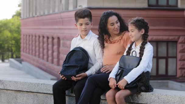 Nuori nainen rakastava äiti äiti opettaja istuu koulun pihalla kadulla ulkona lasten kanssa rakas tytär ja poika koululaiset puhuvat, latino perhe kaksi opiskelijaa ja äiti puhuu koulun jälkeen. - Materiaali, video