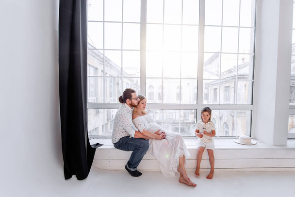 Happy family is having fun while sitting on the windowsill by the panoramic window. Young woman is pregnant, man hugs beloved. Little girl fooling around parents, dancing. Minimalist loft interior - Fotó, kép