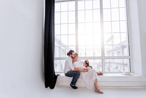 The little girl laughs lies on the lap of pregnant mother the father tickles daughter. Happy family having fun on the windowsill by the panoramic window in white minimalist interior. Healthy pregnancy - Valokuva, kuva