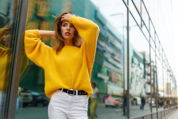 若いです自信女性とともに茶色の波状の髪,現代的な建物の後ろに黄色のセーターと白いジーンズで着用. - 写真・画像