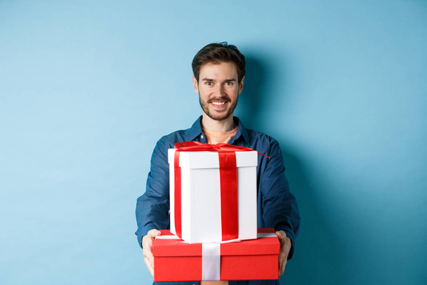 Szczęśliwych walentynek. Przystojny mężczyzna daje dziewczynie prezenty, trzyma pudełka z prezentami i uśmiecha się, stojąc na niebieskim tle - Zdjęcie, obraz