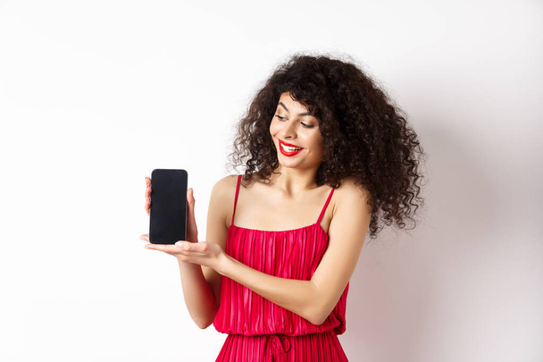 Schönheit Frau mit Make-up und lockigem Haar, zeigt leeren Smartphone-Bildschirm, demonstrieren App, steht auf weißem Hintergrund - Foto, Bild