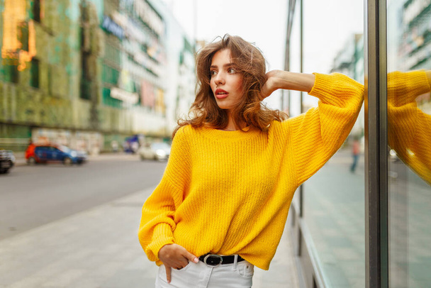 茶色の波状の髪を持つ美しい若い女性は、現代的な建物の後ろに黄色のセーターと白いジーンズを着て. - 写真・画像