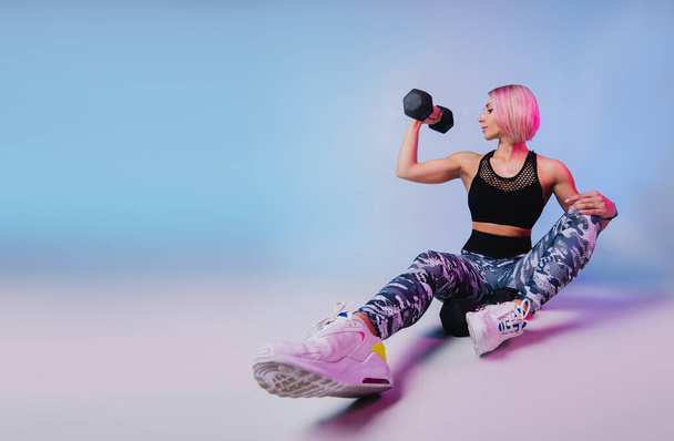 Stylish sports background. Sporty modern woman exercising with dumbbells strong with muscles. Blue fitness neon banner. Workout gym. Girl powerful in sportswear. Bodybuilding. Pink hair  - Foto, afbeelding