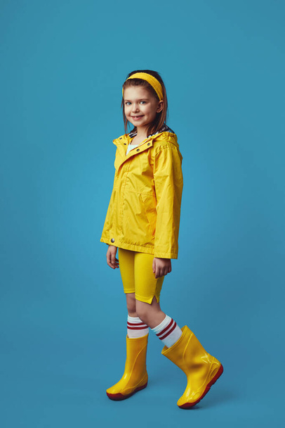 Klein meisje in gele regenjas en sneakers glimlacht terwijl ze tegen blauw staat - Foto, afbeelding
