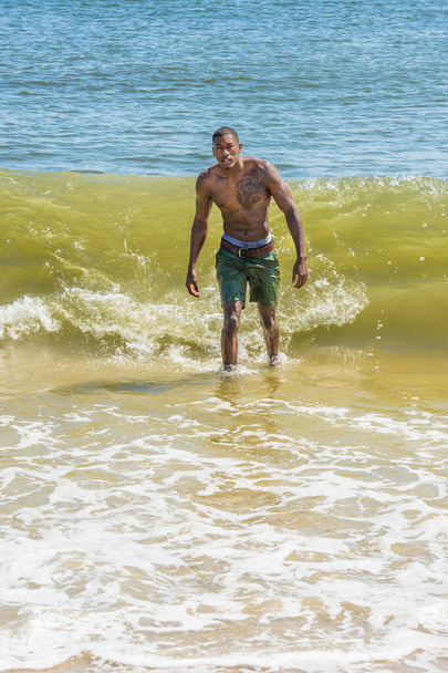 A well built young sexy black guy, half naked,  is confidently walking out off ocean. Huge waves coming from back - Zdjęcie, obraz