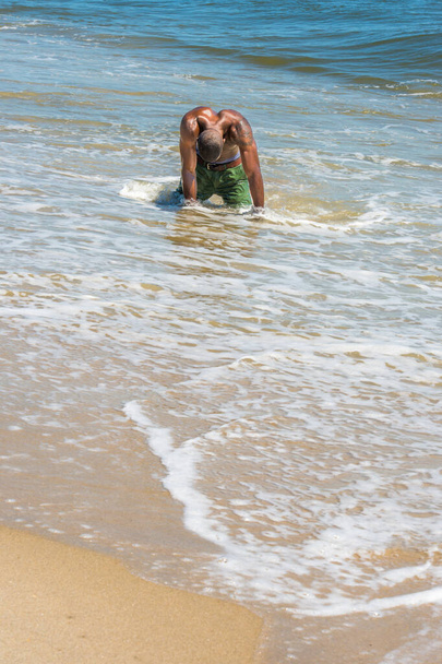 A young black guy, half naked, whole body wet,  is kneeling down on water of the beach, hanging his head, two arm stretching on the ground to support his body - Photo, Image