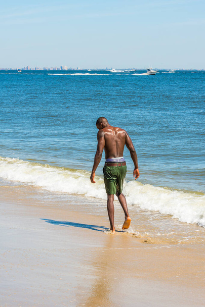 Kun katselee alastomana, koko vartalo märkänä, nuori musta mies kävelee rannalla. Horisontaalinen on suuri kaupungin ääriviivat, pienet veneet taustalla - Valokuva, kuva
