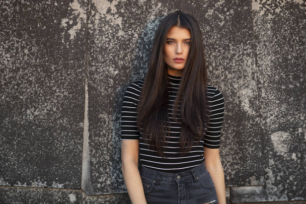 Young girl with straight brown hair, makeup, dressed in modern clothes, supported by a black wall, looking at camera. - Photo, Image